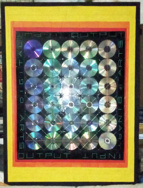 A grid of some thirty iridescent CDs on a black panel bordered by the words DIGITAL ARTS · BINARY · INPUT · OUTPUT, in red, orange and yellow painted borders.
